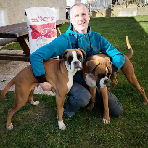 Uomo accovacciato con due cani Boxer e sullo sfondo sacco di crocchette Dogbauer